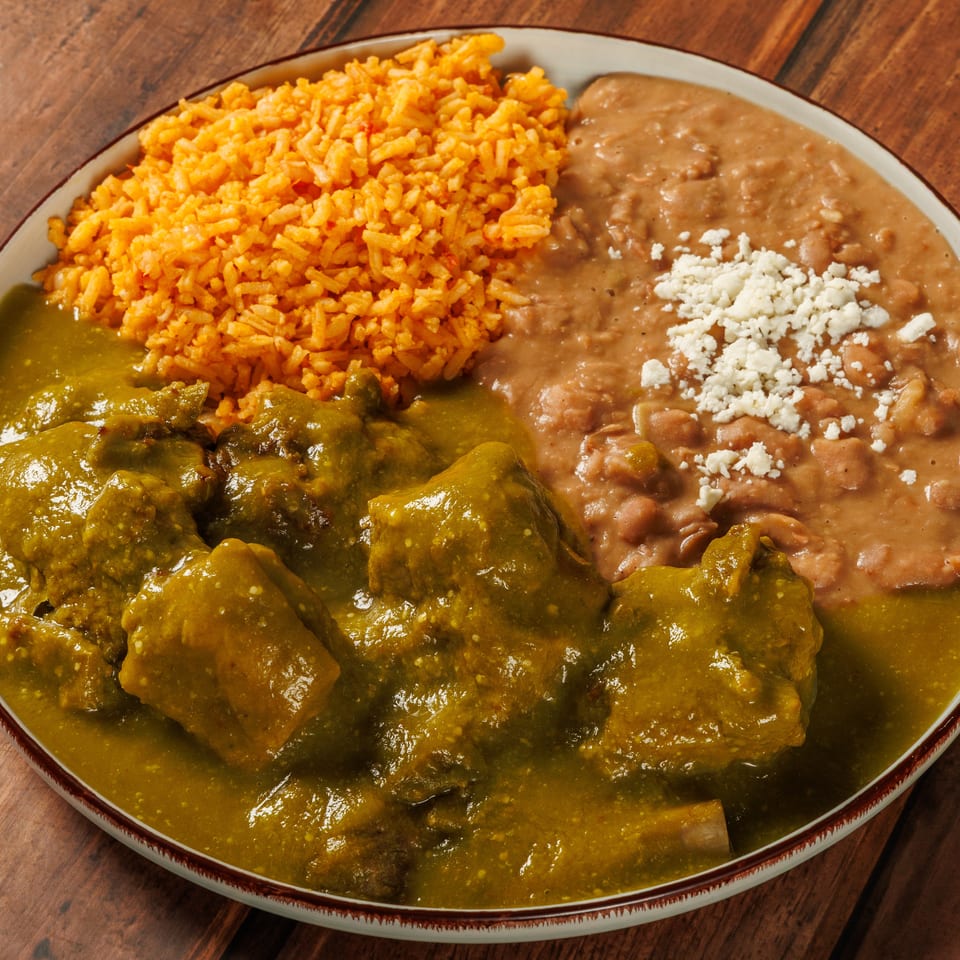 Mexican platter with saucy meat, orange rice, refried beans topped with crumbly cheese, and green chile sauce on a wooden table.