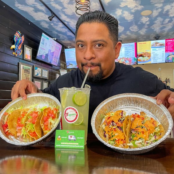 Man holding two foil containers of Taqueros Mexican tacos and sipping a lime drink.