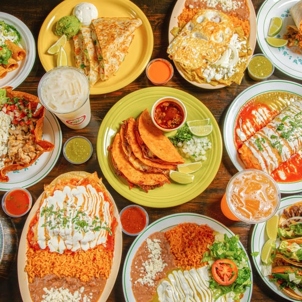 A vibrant spread of Taqueros Mexican dishes, including tacos, quesadillas, burritos, and drinks on a wooden table.