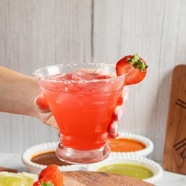 Hand holding a strawberry margarita with a salted rim and strawberry garnish, with sauces in the background.