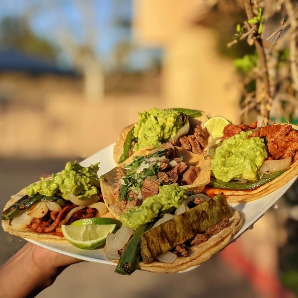 A hand holds a white plate with six Taqueros Mexican Restaurant tacos, topped with guacamole, meat, and lime.