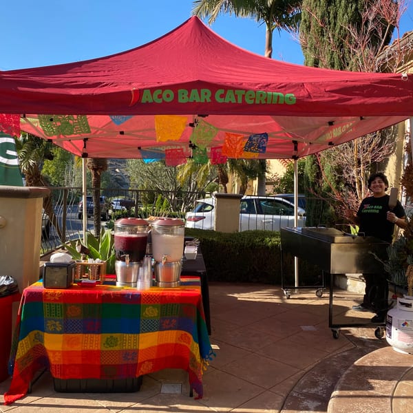 Taqueros Mexican Restaurant catering setup with a red tent, colorful tablecloth, drinks, and a chef grilling.
