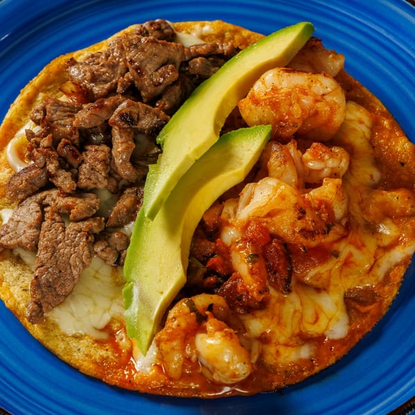 A Taqueros Mexican Restaurant tostada with steak, shrimp, melted cheese, and avocado slices on a blue plate.
