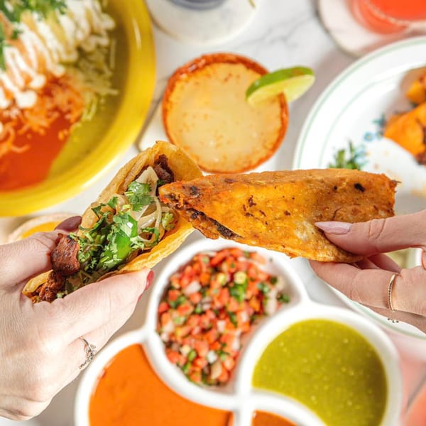 Two hands holding birria tacos over a table with various Mexican dishes and salsas.