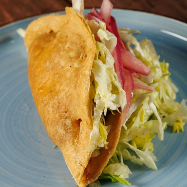 A crispy Taqueros Mexican Restaurant taco filled with shredded lettuce and pickled red onions on a blue plate.