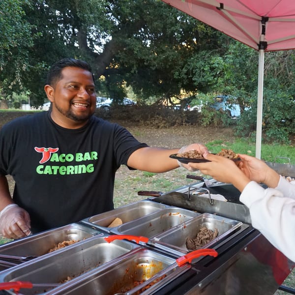 A smiling Taqueros catering staff member serves tacos from a food stall outdoors.
