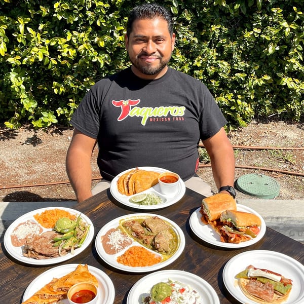 A man in a Taqueros Mexican Restaurant shirt sits at a table laden with various Mexican dishes.