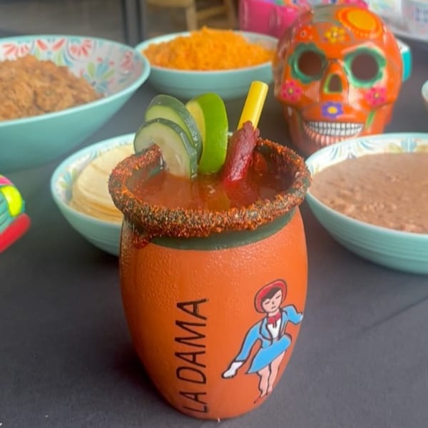A michelada in a clay cup with cucumber and lime, surrounded by Mexican food and a sugar skull.