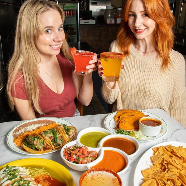 Two women toasting colorful margaritas at Taqueros Mexican Restaurant, surrounded by various Mexican dishes.