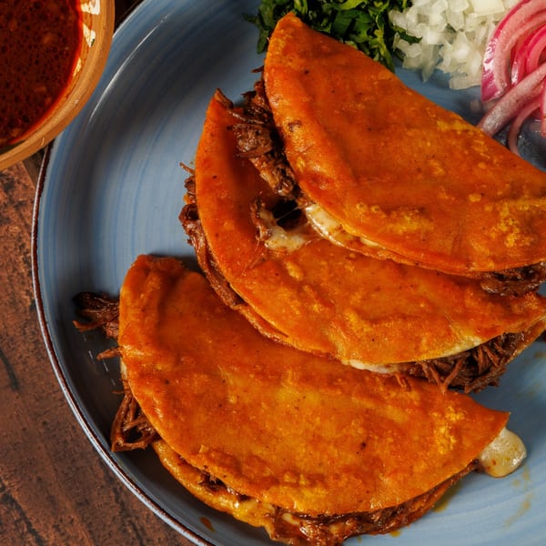 Three cheesy birria tacos with shredded meat, served with a side of consommé, onions, and cilantro.
