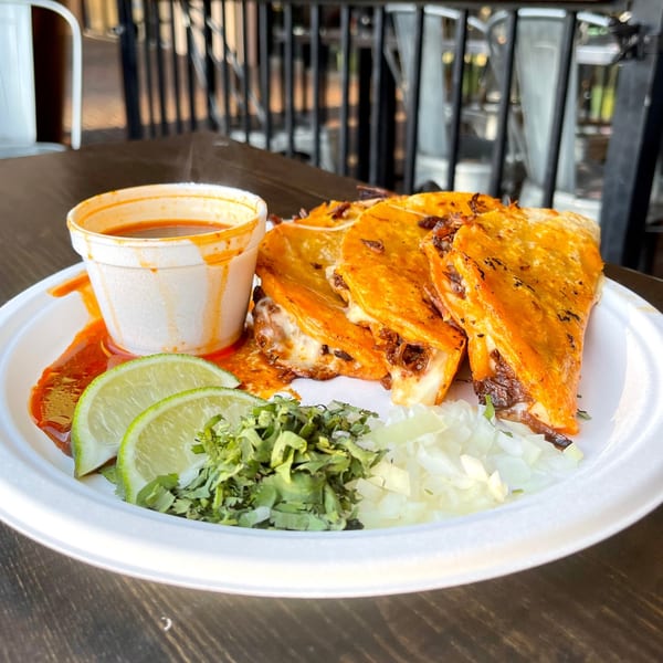 Three cheesy birria tacos with a side of consommé, lime wedges, chopped cilantro, and onions.