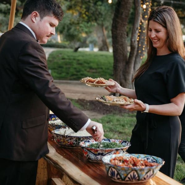 A man and woman serving tacos and toppings from a buffet line outdoors at Taqueros Mexican Restaurant.