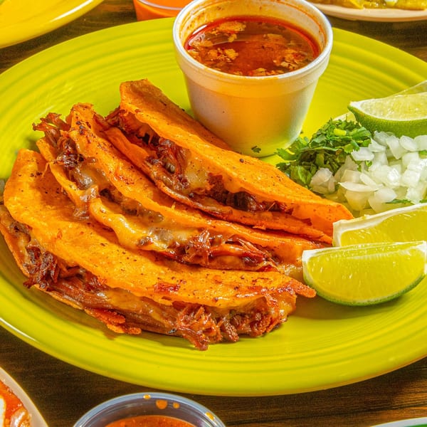 Birria tacos with consommé, cilantro, diced onions, and lime wedges on a bright green plate.