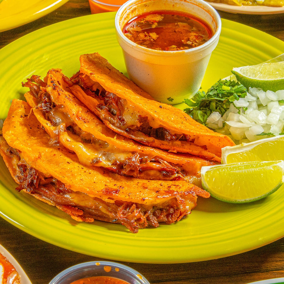 Birria tacos with consommé, cilantro, diced onions, and lime wedges on a bright green plate.