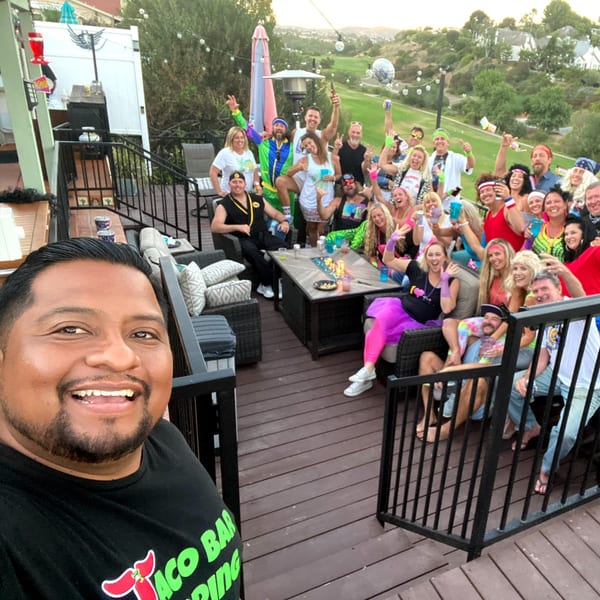 A man takes a selfie with a large group of people celebrating on an outdoor patio.