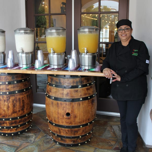A Taqueros catering staff member smiles next to a beverage station with horchata and orange juice.